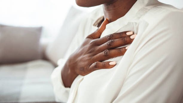 Girl having respiration problems touching chest sitting on a couch in the living room at home.  Woman feeling pain ache touching chest having heart attack, sad worried lady suffers from heartache at home.