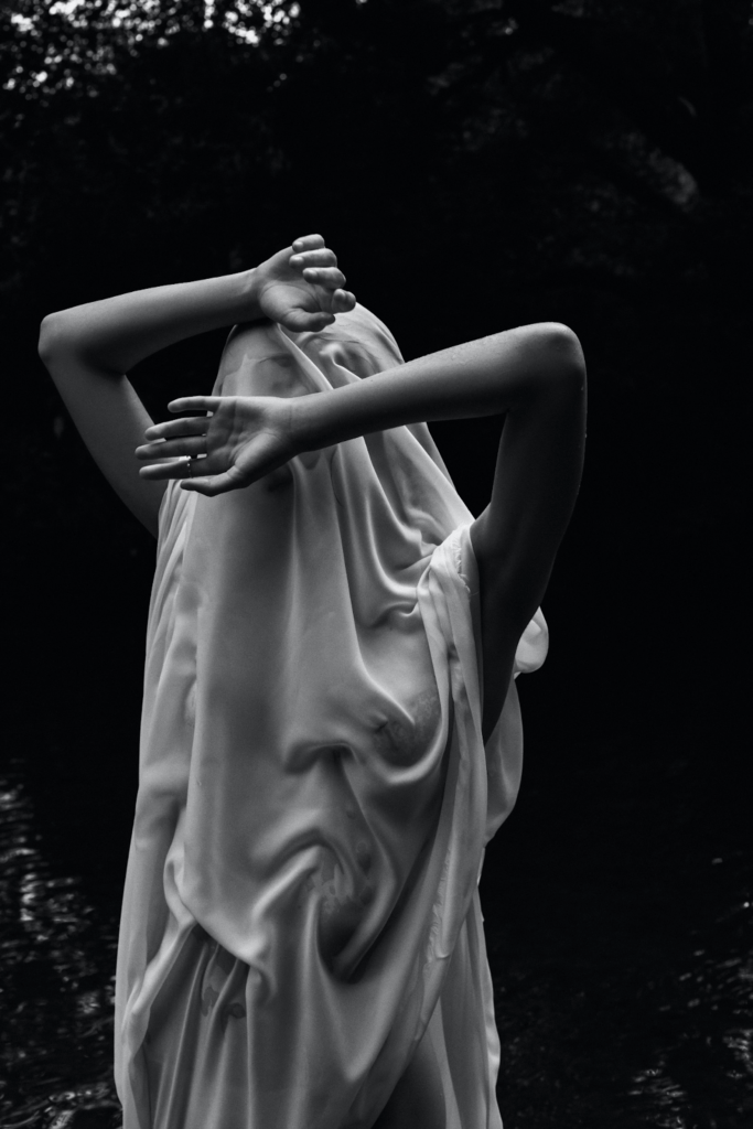 a black and white photo of a woman covered in a white sheet with her arms up near her face