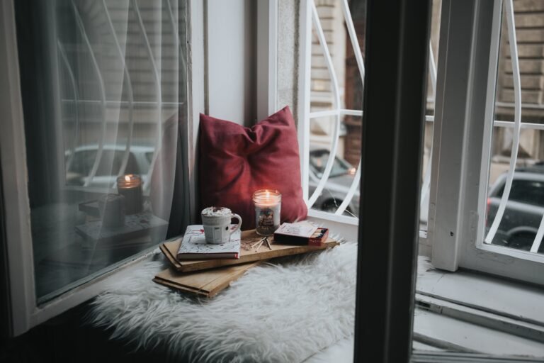 Candle, cup of hot chocolate, book, and pillow stacked in a window nook