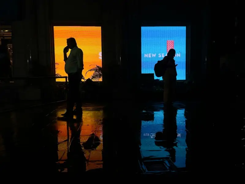 Two people stand in shadow, one in front of a blue lit up sign; the other facing oppositely in front of a yellow lit up sign.